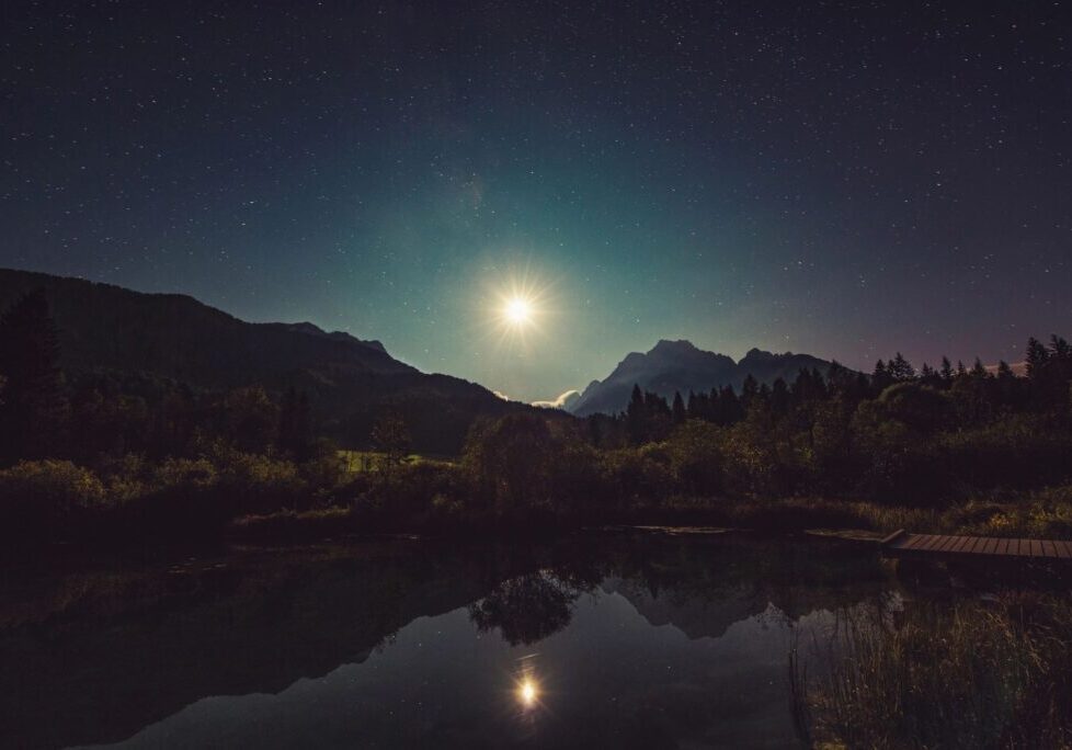 Full moon shining over a calm lake surrounded by mountains at night.